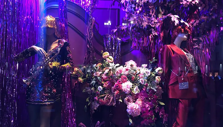 The image shows two mannequins adorned with stylish outfits and unique headpieces, positioned among a vibrant backdrop of metallic streamers and a lush floral arrangement.