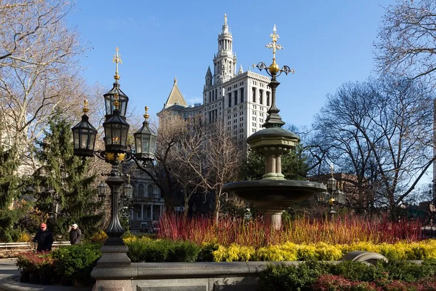 An ornate fountain and vintage street lamps stand amidst colorful plant life in an urban park with a tall, historic building in the background.