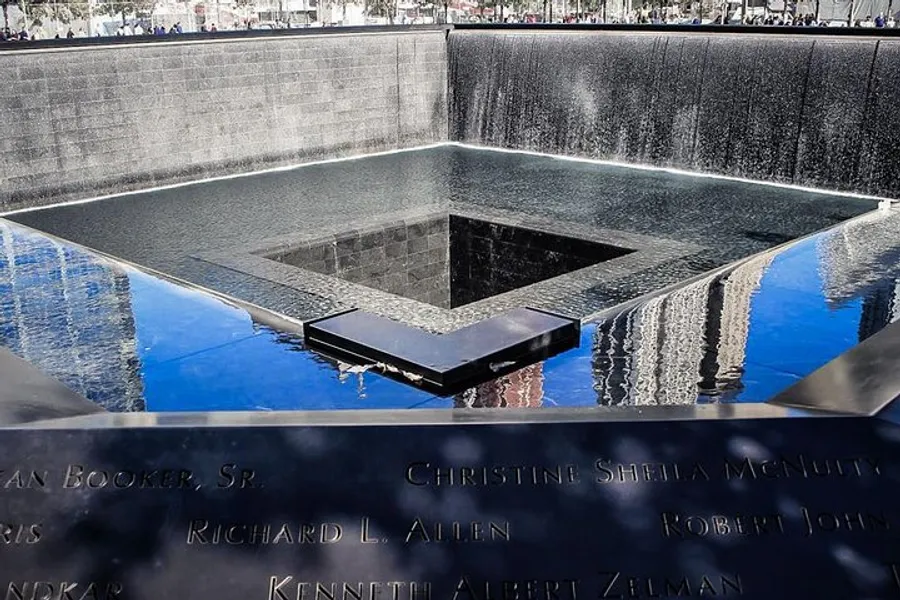 The image depicts a large, square memorial pool with water cascading down its sides, surrounded by panels with inscribed names, as part of a somber commemorative site.