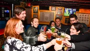 A group of cheerful people are toasting with drinks in a cozy bar with sports playing on a TV in the background.