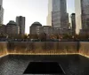 The image shows a serene night view of the 911 Memorial featuring one of the twin reflecting pools with illuminated skyscrapers rising in the background