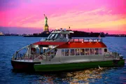 A ferry boat is cruising on the water with passengers on board, against a backdrop of a pink-hued sky with the Statue of Liberty in the distance.