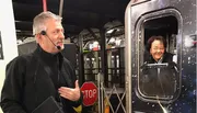 A man with a headset is speaking and gesturing while a woman smiles from the open doorway of a subway car, with a 