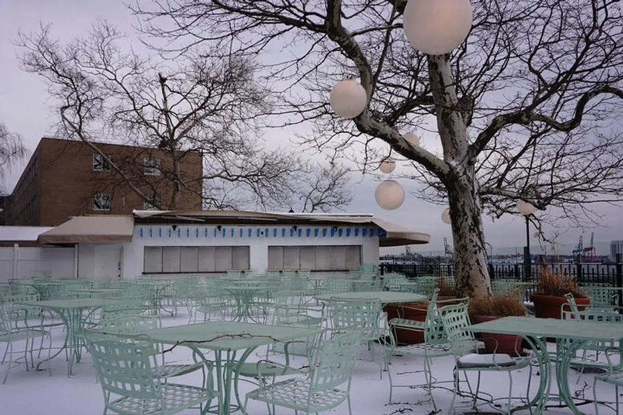 An outdoor seating area with green metal chairs and tables is covered in snow, framed by a leafless tree and spherical light fixtures, with a building and overcast sky in the background.
