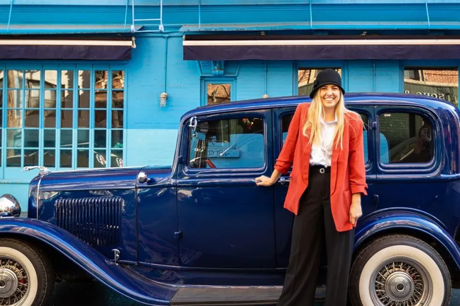 A smiling person in a red jacket and black hat stands next to a classic blue car in front of a building with a bright blue facade.