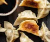 The image shows several golden-brown pan-fried dumplings next to a small bowl of dipping sauce on a dark surface