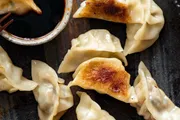 The image shows several golden-brown pan-fried dumplings next to a small bowl of dipping sauce on a dark surface.