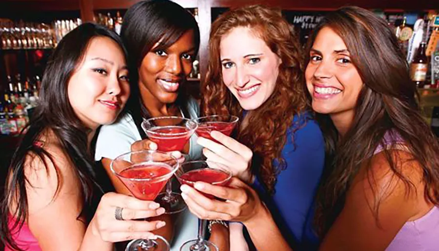 Four smiling women are holding and toasting with red cocktails at a bar.