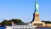 A ferry full of passengers sails near the Statue of Liberty under a clear blue sky.