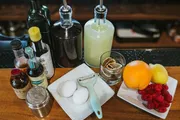 The image displays various cocktail ingredients and tools, including citrus fruits, berries, eggs, bitters, syrups, and a juicer, arranged on a bar counter.