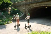 Two people wearing helmets are enjoying a ride on electric scooters near a historical archway in a park setting.