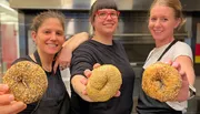 Three smiling people are holding up large, seeded bagels toward the camera in a kitchen setting.