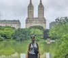 A person is standing in front of a serene pond with towering buildings in the background likely in a city park