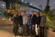 A group of smiling people pose for a photo at night with a lit-up bridge and city skyline in the background.