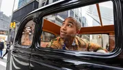 A person is leaning out the window of a vintage car, looking up with a look of wonder or excitement, set against an urban street backdrop.