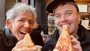 Two individuals are smiling at the camera while each biting into large slices of pizza.