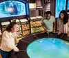 Three people are interacting with a large circular interactive display possibly a digital exhibit in a room that features shelves of wand boxes and screens showing images related to the Harry Potter series