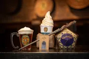 The image features an assortment of Harry Potter-themed items including a butterbeer in a mug with whipped cream on top, a soft-serve ice cream cone, a chocolate frog package, a wand, and a brown paper bag with a logo, all set against the backdrop of barrels.