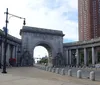 The image shows a large intricately carved triumphal arch flanked by colonnades situated at the entrance of a plaza with a city background and a high-rise building to the right