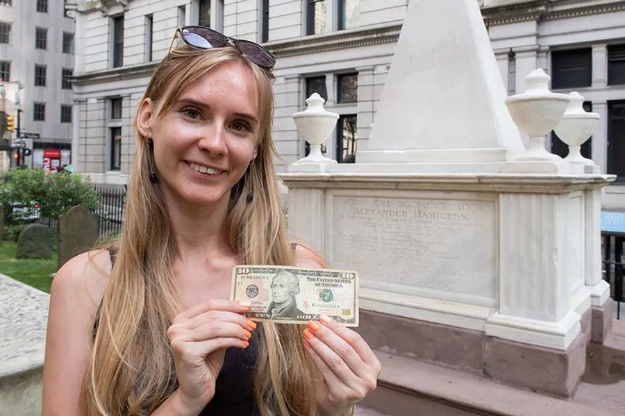 A smiling woman is holding a ten-dollar bill in front of the Alexander Hamilton memorial.