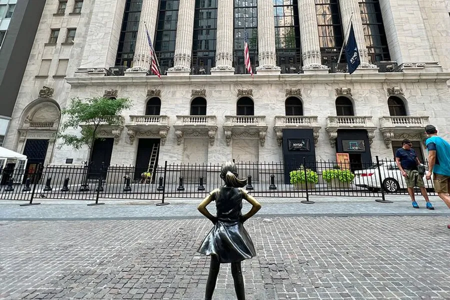 A bronze statue of a girl stands with her hands on her hips facing the New York Stock Exchange, with passersby and American flags in the background.
