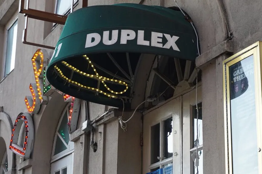 The image shows an outdoor awning with the word DUPLEX above a door, adorned with a lit neon sign shaped like a cocktail glass.
