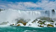The image shows the powerful and picturesque Niagara Falls with cascades of water plunging over a crest lined with white frothy water and mist, backed by a clear blue sky speckled with clouds.