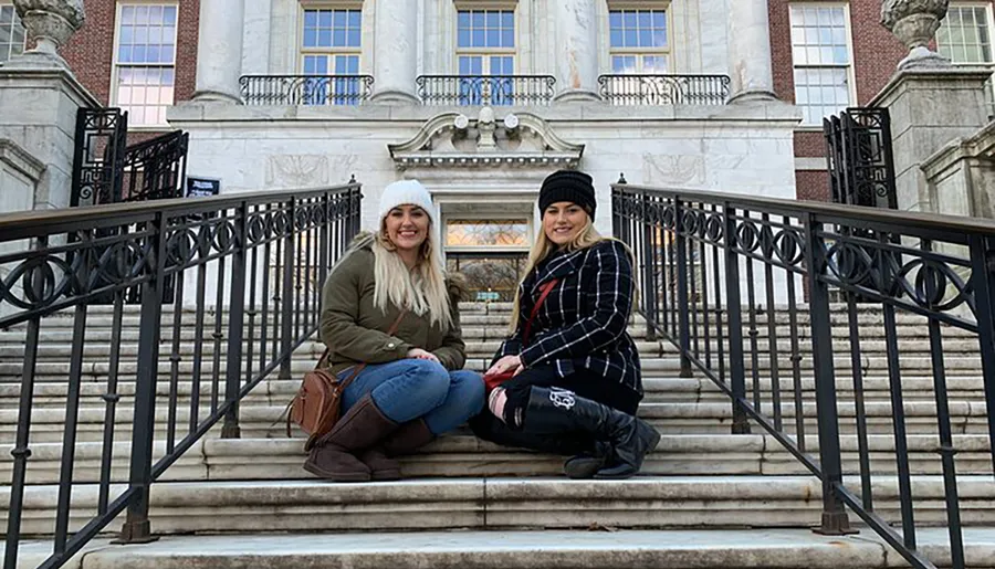 Two people are sitting on the steps in front of an ornate building with a classic façade and smiling at the camera.