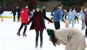 A group of people are enjoying ice skating, with a smiling woman in the center balancing herself while another person appears to be bending down or falling in the foreground.