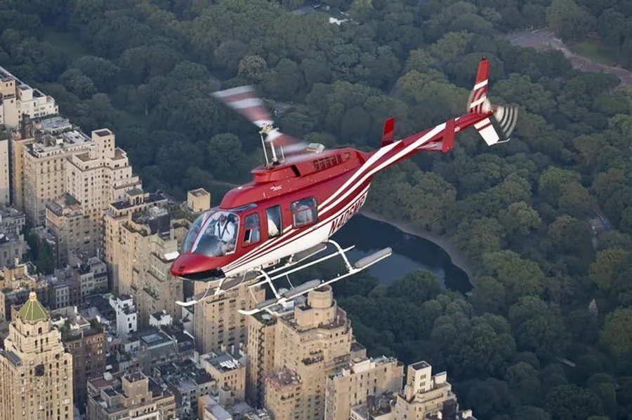 A red and white helicopter is flying over an urban area adjacent to a large, park-like green space.