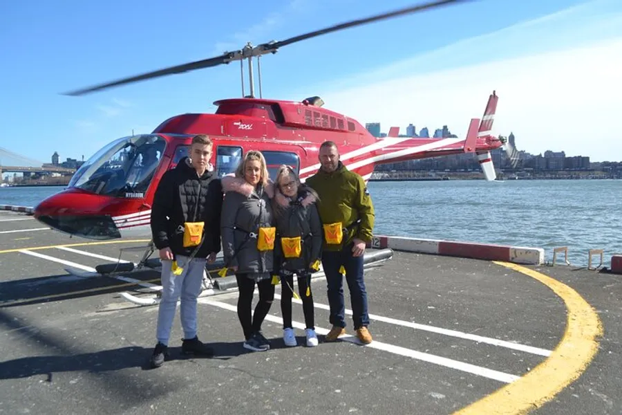 Four people are smiling for a photo in front of a red helicopter on a helipad near a body of water, with a city skyline in the background.