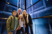 A family is looking up with expressions of awe and excitement while standing in what appears to be a glass elevator or enclosure.
