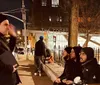 A group of people is sitting and standing on a city sidewalk at night engaging in conversation near a building labeled THE COOPER UNION