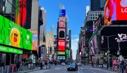 The image captures the vibrant atmosphere of Times Square in New York City, bustling with pedestrians and adorned with iconic illuminated billboards under a clear blue sky.