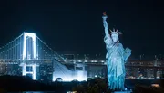 The image captures a nighttime scene of a Statue of Liberty replica with a brightly illuminated suspension bridge in the background.