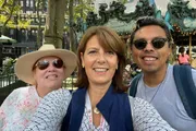 Three people are smiling for a selfie in front of a carousel in a bustling outdoor area dense with trees and street furniture.