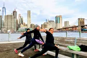 Three people are performing yoga poses on a waterfront with a city skyline and a bridge in the background.
