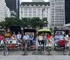 Three people are sitting in a pedal-powered rickshaw enjoying a sunny day in a park with other rickshaws and visitors in the background