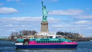 A colorful tourist boat sails near the Statue of Liberty under a blue sky with streaky clouds.