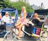 Three passengers are enjoying a ride in a pedal-powered rickshaw while a photographer captures the scene in a park with snow patches on the ground and a lake in the background