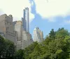 The image shows a view of Central Park with lush greenery in the foreground and a skyline of mixed architectural styles under a partly cloudy sky