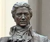 The image shows a close-up of a bronze statue of a historical figure set against a blurred background with architectural elements