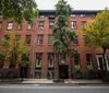 The image shows a row of red-brick townhouses with large windows one of which is covered with green ivy situated on a tree-lined urban street