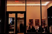 The image captures a warm and inviting view into a restaurant at night, seen from the outside through large windows, where people are dining and conversing under the soft interior lighting with colorful artwork on the walls.