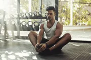 A person is stretching on the floor of a gym with weights in the background, bathed in natural light coming through large windows.