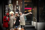 A group of cheerful people pose for a photo on a city sidewalk, with one mimicking the mannequin's pose in the background.