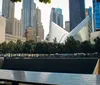 The image shows the 911 Memorial with its reflecting pools in the foreground and the distinctive white ribs of the Oculus structure in the background within a modern urban skyline