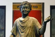 This image shows a bronze sculpture of a young boy in classical style, raising his right hand possibly in gesture, with a smiling expression and curly hair, set against a blurred background with red and black elements.