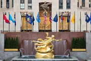 The image shows the iconic golden Prometheus statue overlooking the ice skating rink at Rockefeller Center, against a backdrop of international flags and the bold artwork above the rink.