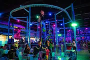 The image depicts an indoor amusement center with vibrant lighting, featuring a roller coaster track above, various play structures, and people seated and walking around.